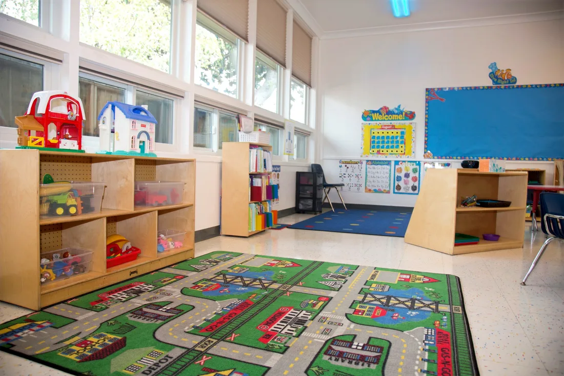 A classroom with open shelves of dollhouses and toy vehicles beneath a wall of windows, and a large road rug across the floor.