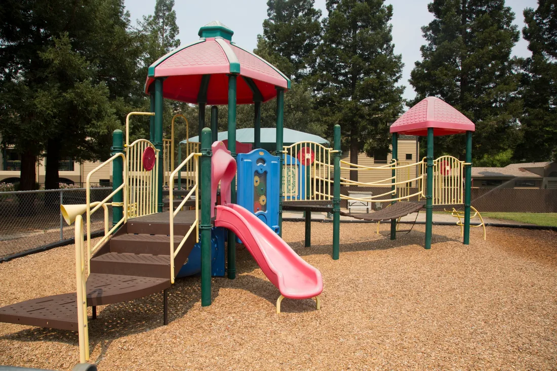 The main play structure with its slide and two shaded towers, on bark chips, with tall redwoods behind it.